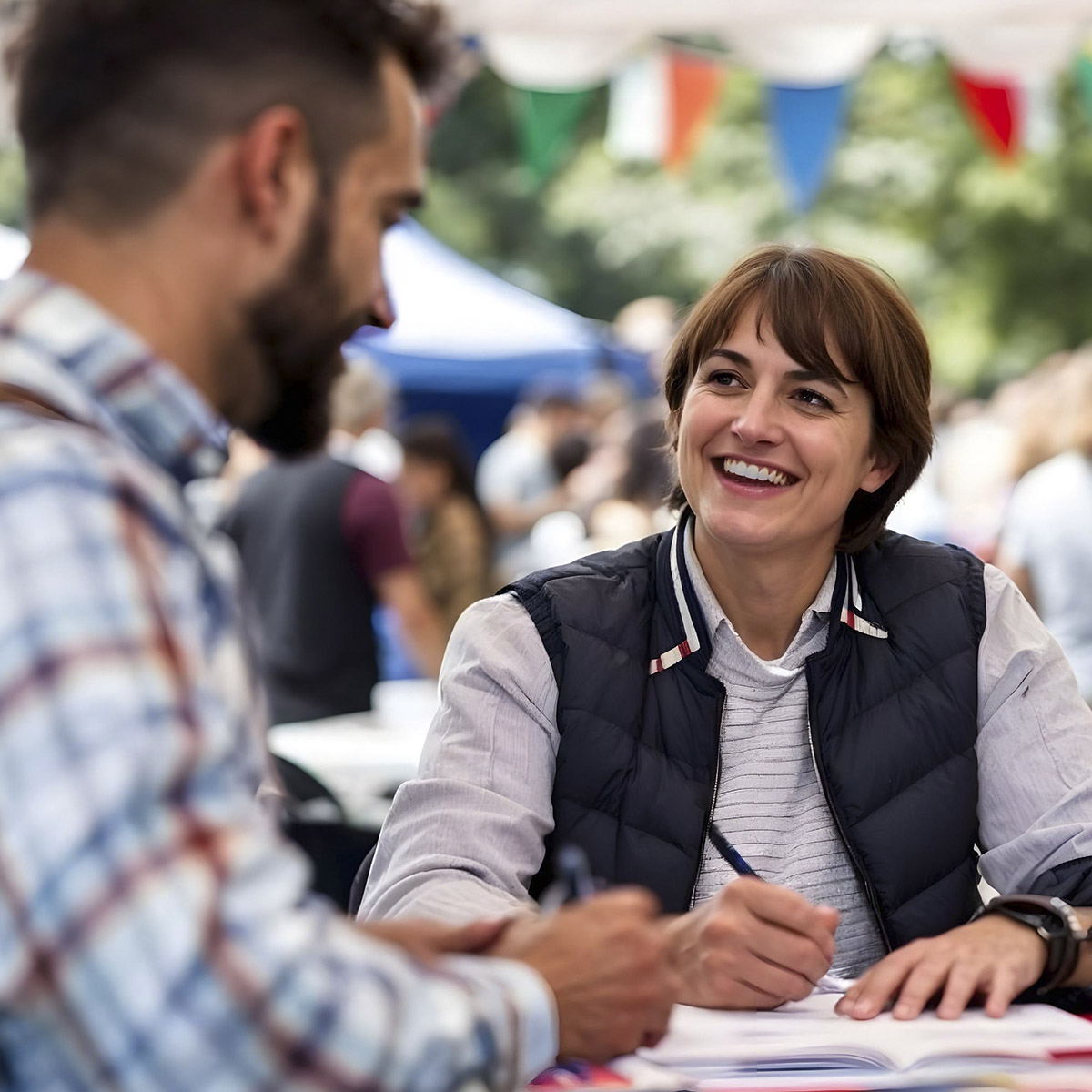 writers-scene-concept_squarecrop A woman in a vest smiles while writing as she talks to a man at an outdoor event.