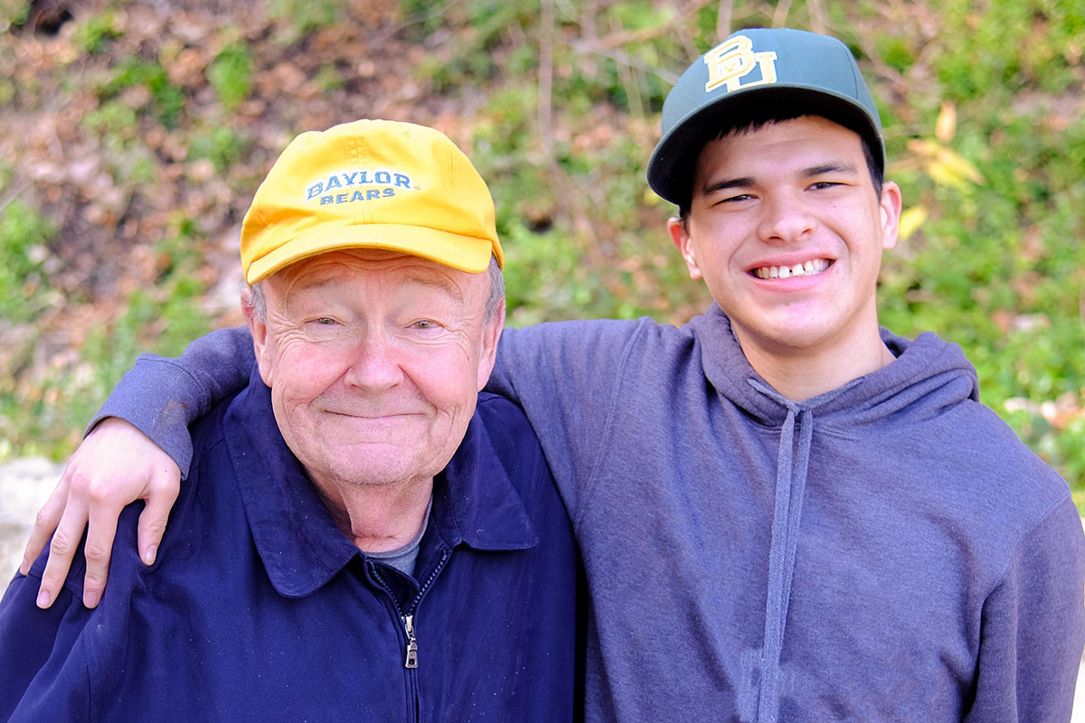 2024.02.12.54 3x2 crop An elderly man in a yellow cap and a young man in a dark hoodie smile warmly, standing outdoors against a leafy background, exuding joy and camaraderie.