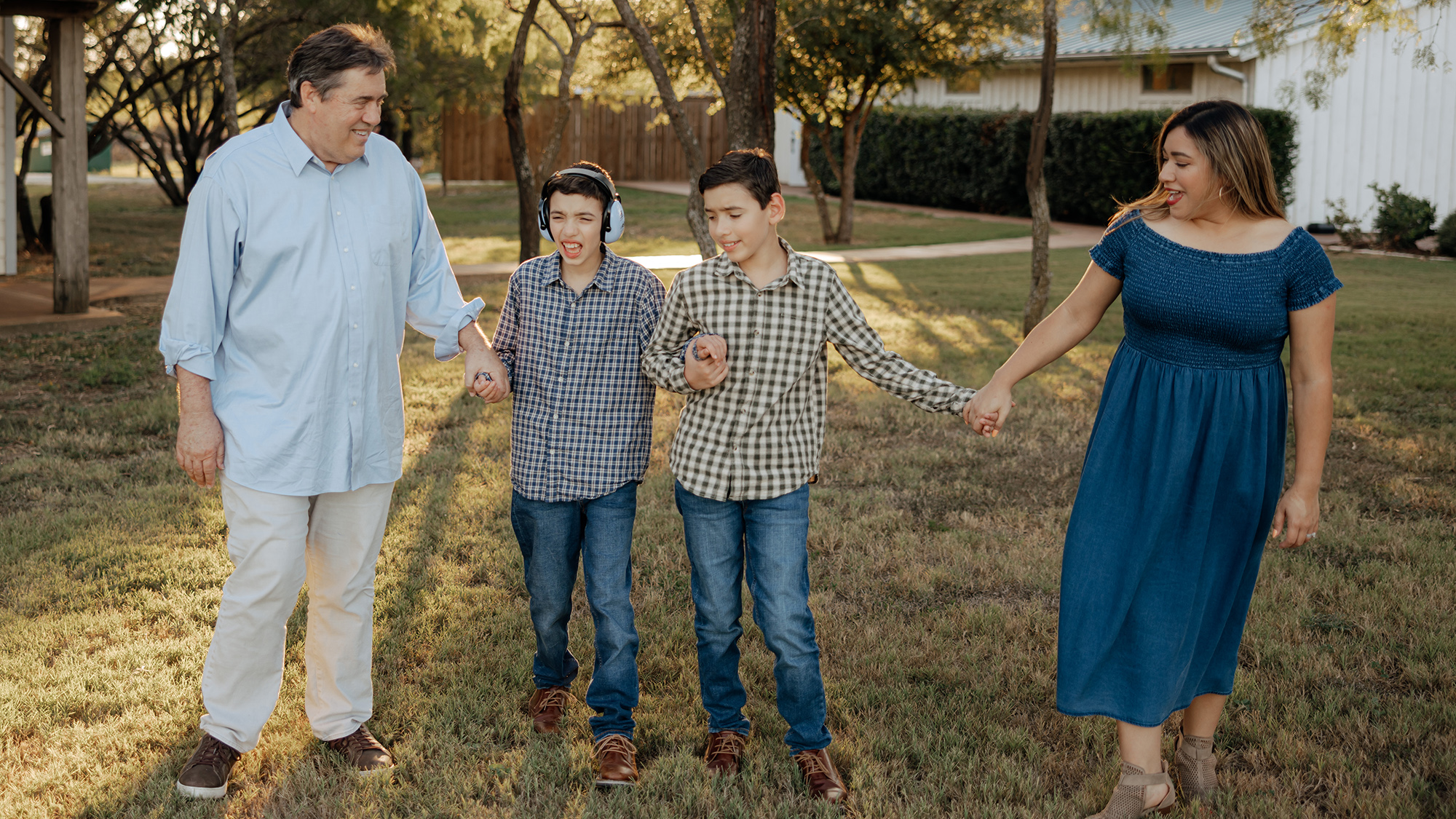 A family walks hand in hand through a grassy yard. Two children, one wearing headphones, are with their parents. The scene conveys warmth and togetherness.