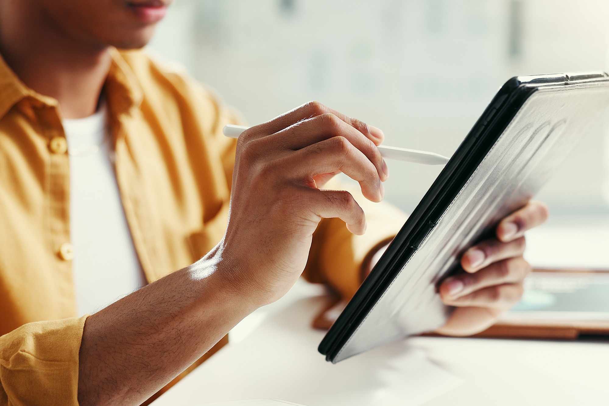 hands-tablet-3x2 crop A person in a mustard-colored shirt uses a stylus to interact with a tablet, conveying focus and concentration in a bright, modern setting.