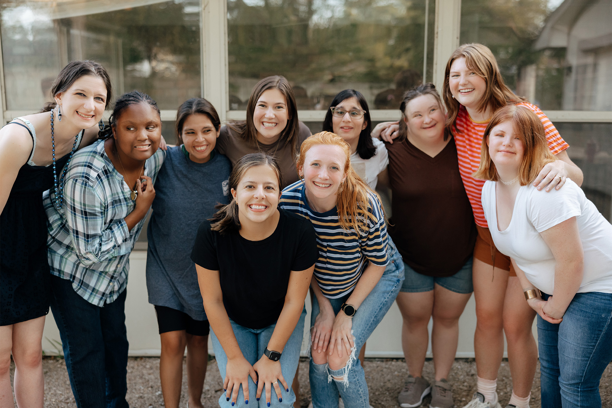 A group of ten smiling young women, diverse in appearance, gather closely. They exude joy and camaraderie, posing casually in front of a window.