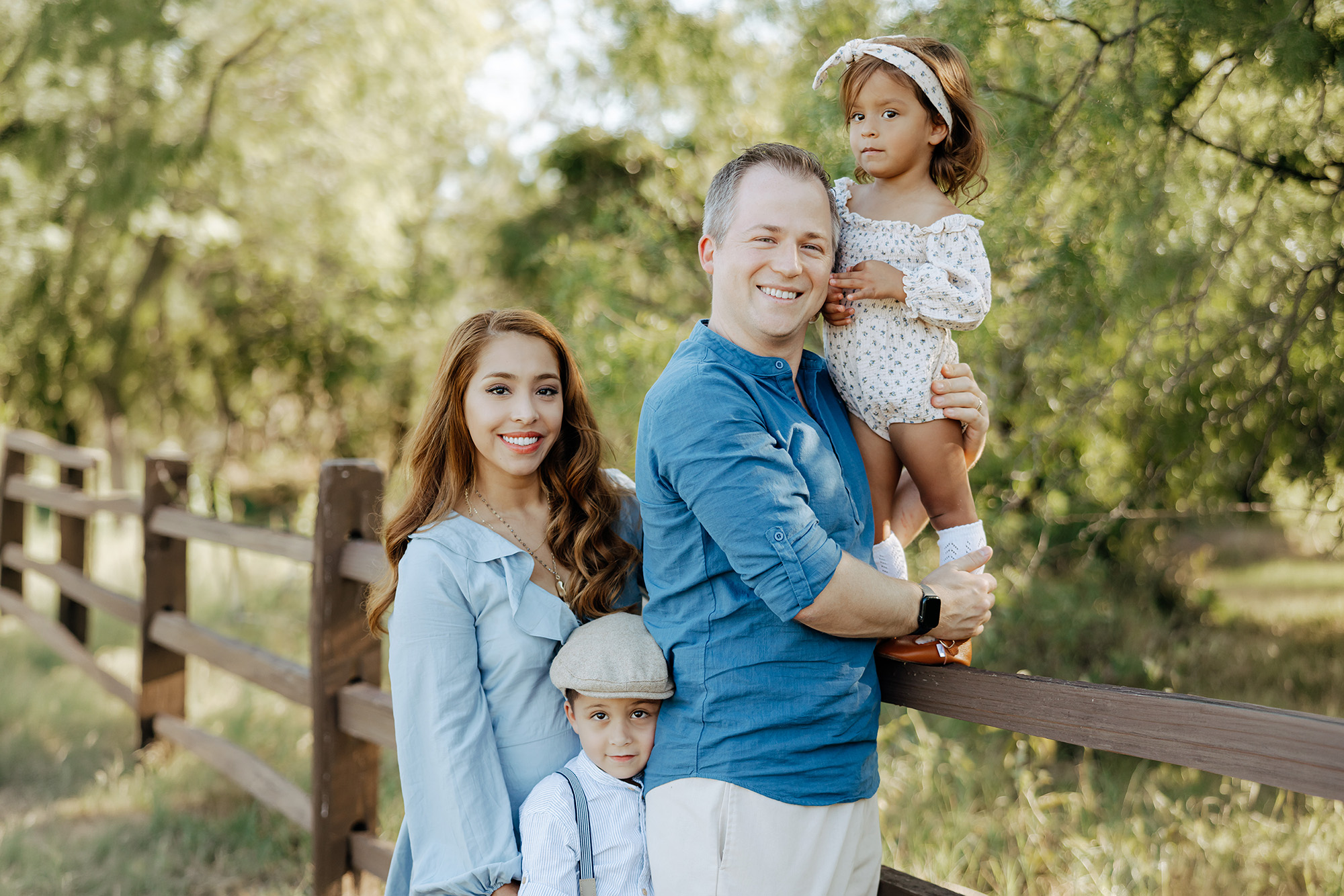 A family of four poses outdoors by a wooden fence, with trees in the background. The father holds a young girl, while the mother stands beside a young boy in a cap.