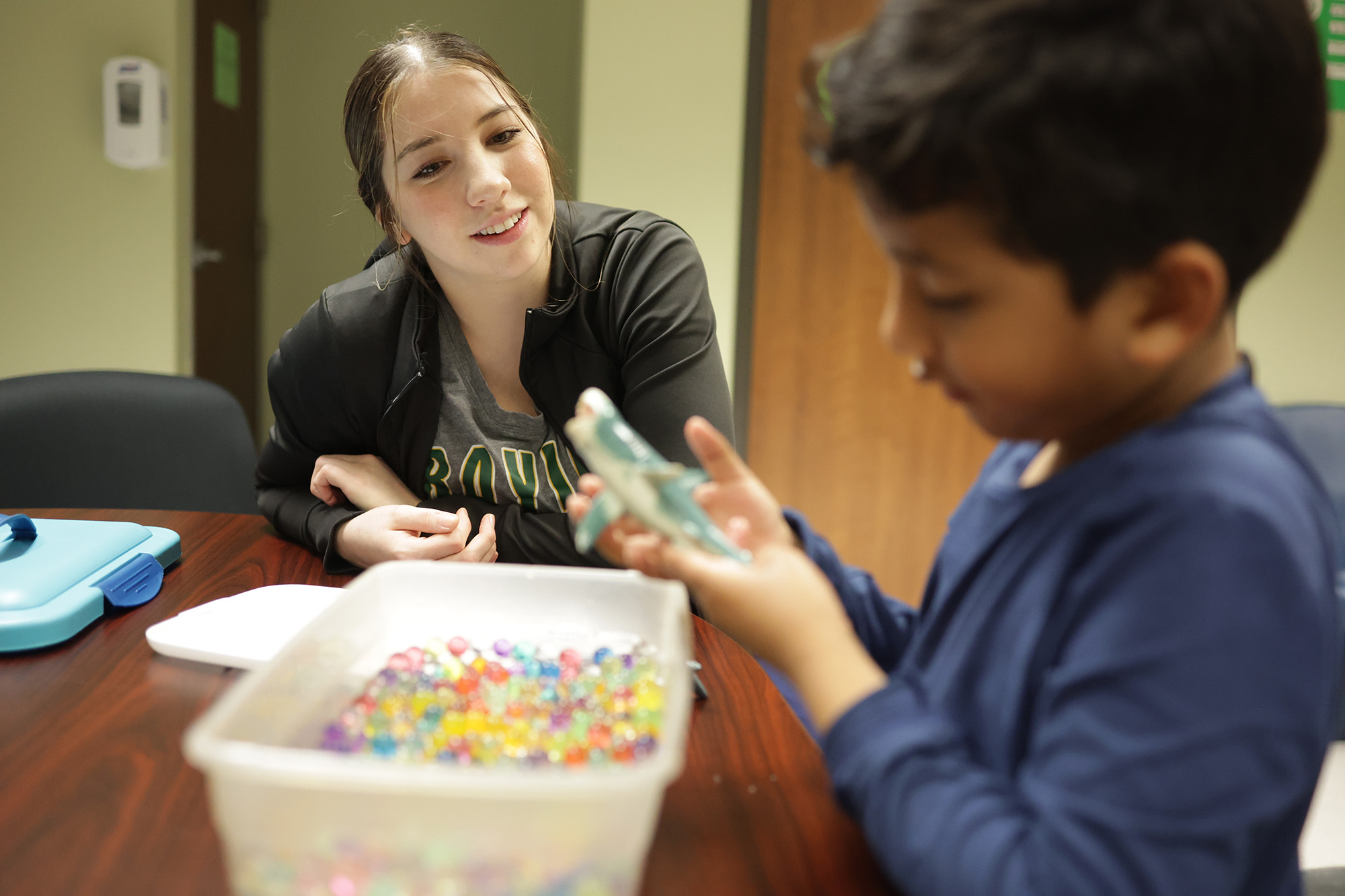 A woman smiles at a boy holding a toy while sitting at a table with colorful beads. The scene conveys a cheerful and focused learning environment.