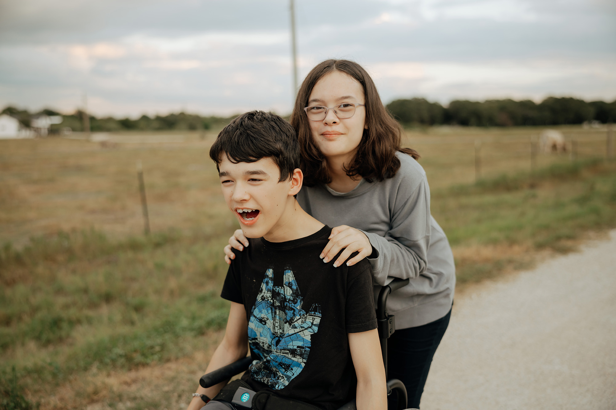 A boy in a wheelchair smiles while a girl stands behind him with her hands on his shoulders. They are outdoors on a rural path with fields in the background.